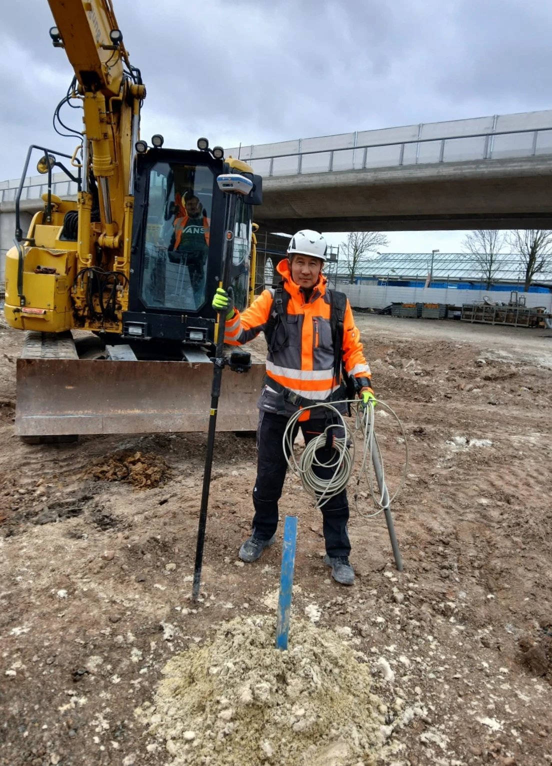 Mitarbeiter mit Messausrüstung vor einem gelben Bagger auf der Baustelle.